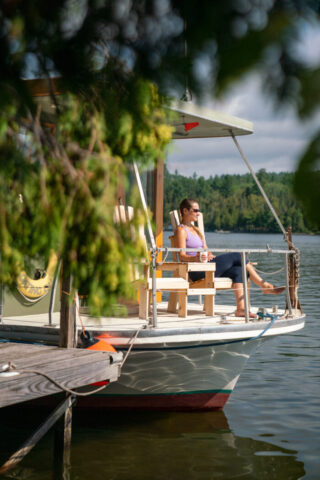 A woman in athletic wear and sunglasses holding a cup of coffee sits on the bow of a houseboat moored to a dock. Captured by photographer Kirsten Alana.